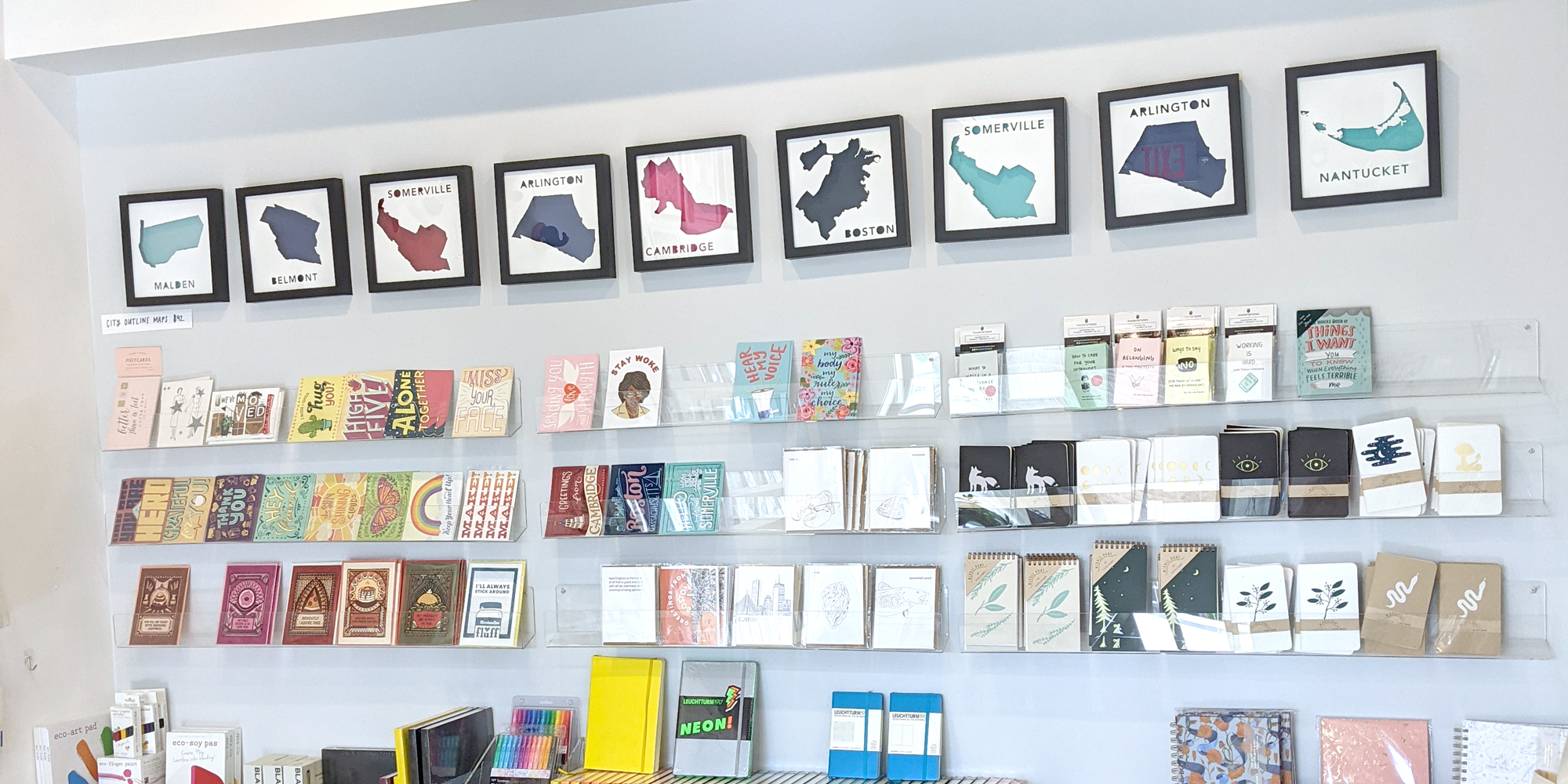A variety of framed paper places on the wall of Tiny Turns Paperie in Somerville, MA above a display of postcards and notebooks