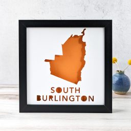 A map of South Burlington, Vermont cut out of white paper in a black frame with an orange background
