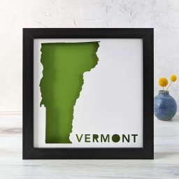 A map of the state of Vermont cut out of white paper in a black frame with a green background