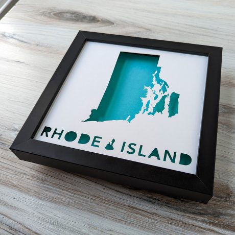 Teal Rhode Island map in black frame on white background, frame is resting on a woodgrain background