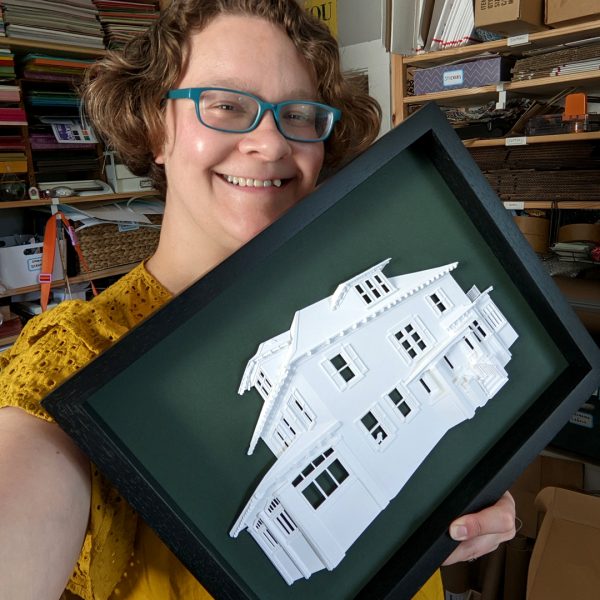 Abigail holding a finished portrait of a two-story home with a silhouette of a small dog in one window. The house is on a dark forest green background in a black frame.