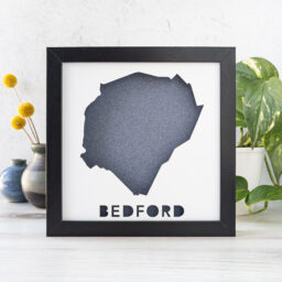 A dark blue and white map of Bedford, MA in a black frame on a desk with a plant and a vase