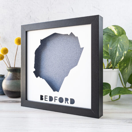 a square black frame with the shape of Bedford, MA cut out of a white piece of paper to reveal a denim blue paper