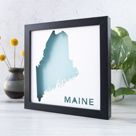 A framed map of the state of Main with the shape of the state cut out from white paper to reveal a light blue background