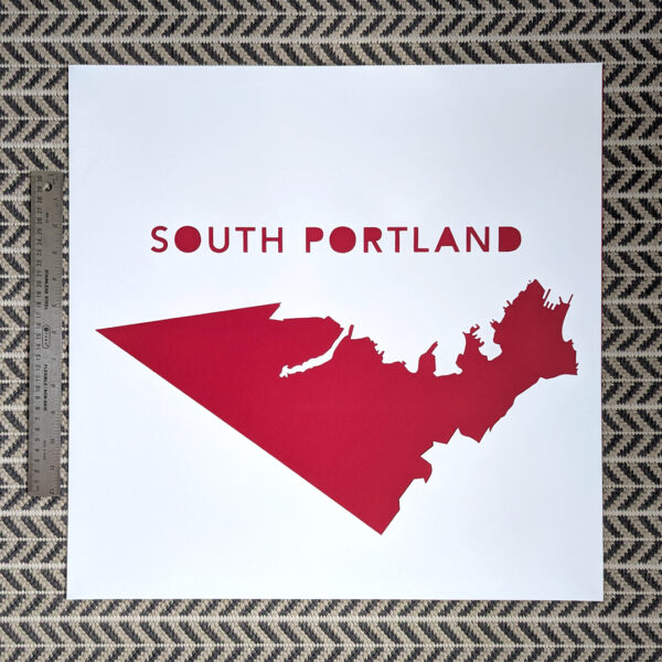 a piece of white paper with a map of south portland, Maine cut out to reveal a red background, with a ruler for scale along the left side