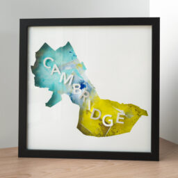 A framed map of the city of Cambridge, MA cut out of white paper with a blue and yellow-green collage below, with 3d letters spelling "Cambridge"