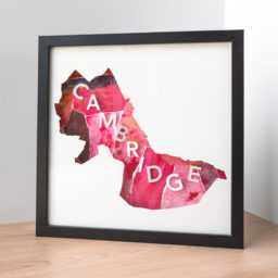 A framed map of Cambridge, MA, the shape of the city is cut from white paper to reveal a pink and red collage with the word "Cambridge" spelled out of 3D paper letters