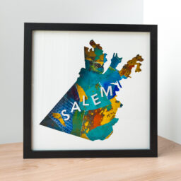 A framed map of Salem, MA. The shape of the city is cut from white paper to reveal a blue and yellow collaged background with 3D paper letters that spell "Salem"
