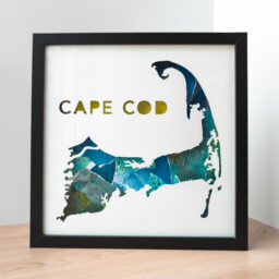 A framed map of Cape Cod, MA. The shape of the peninsula and the words "Cape Cod" are cut from white paper to reveal a textured blue and green collaged background
