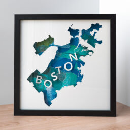 A framed map of Boston, MA. The shape of the city is cut from white paper to reveal a collaged blue and green background with 3D paper letters that spell "Boston"