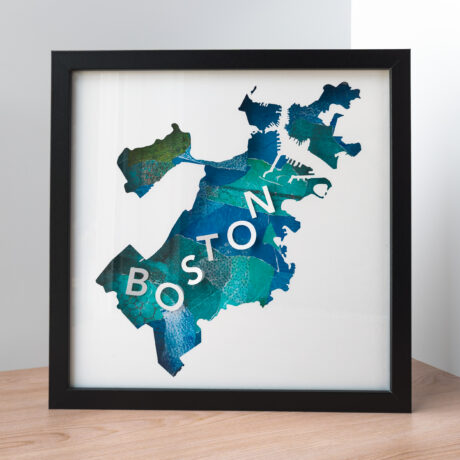 A framed map of Boston, MA. The shape of the city is cut from white paper to reveal a collaged blue and green background with 3D paper letters that spell "Boston"