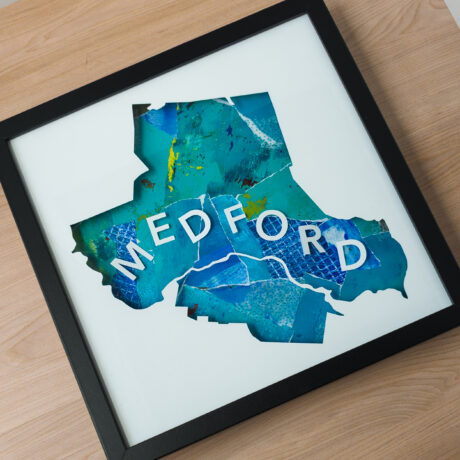 A square framed map of Medford, MA with a blue collaged background laying on a wooden table