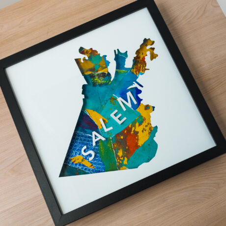 A square framed map of Salem, MA with a textured blue, green, and yellow background. The map is laying on a wooden table.