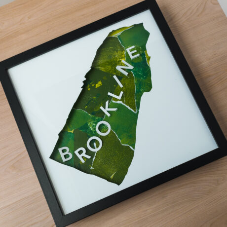 A framed map of Brookline, MA with a green background laying on a wooden table