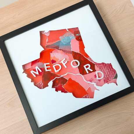 A square framed map of Medford, MA with a red ad pink background laying on a wooden table.