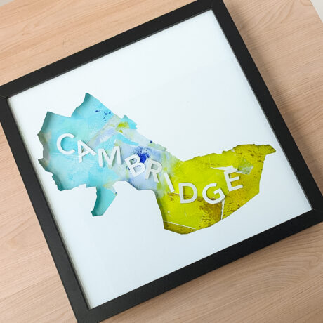 A square framed map of Cambridge, MA with a blue and yellow-green background collage sitting on a wooden table.