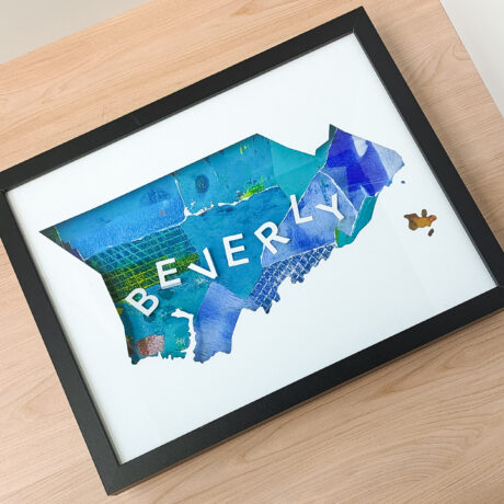 A rectangular framed map of Beverly, MA with a blue and green collaged background laying on a wooden table.